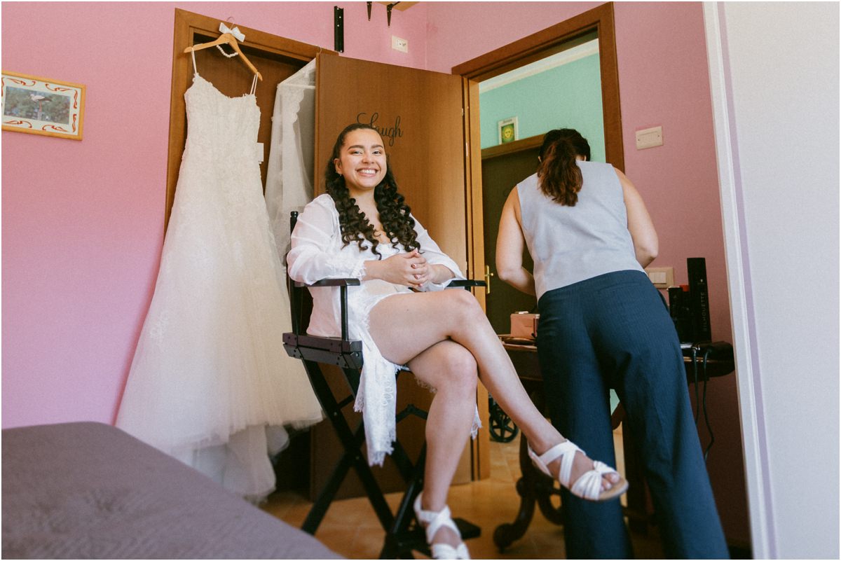 La sposa ride seduta sulla sedia del trucco con l'abito da sposa appeso alla porta