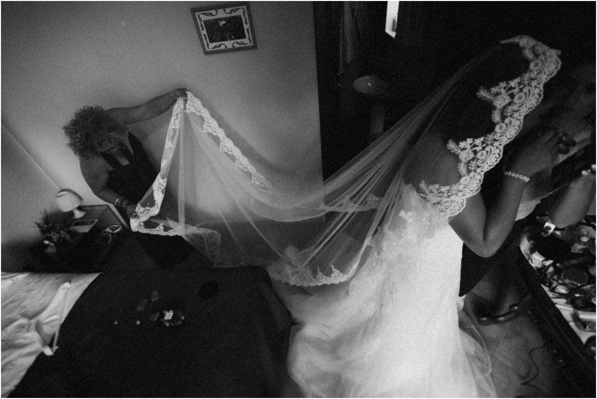 Foto in bianco e nero della mamma che distende il lungo velo della sposa nella camera