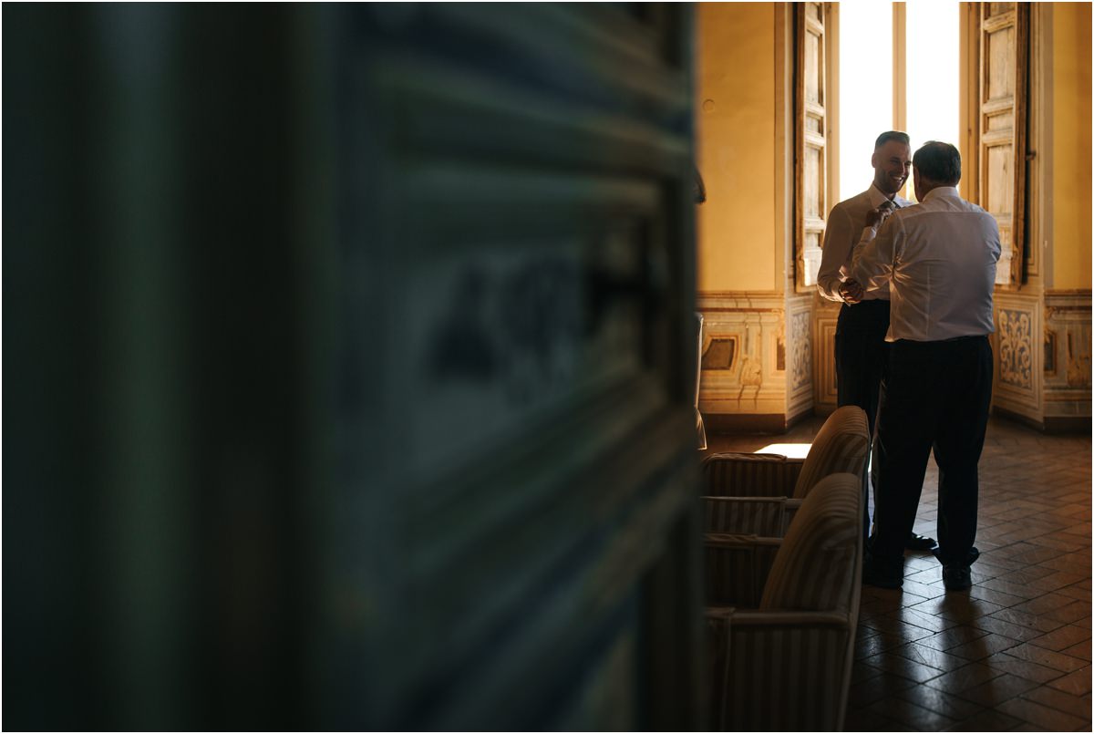 Groom Preparations by the Window Groom and a family member standing by a tall window in a historic room of Villa Grazioli