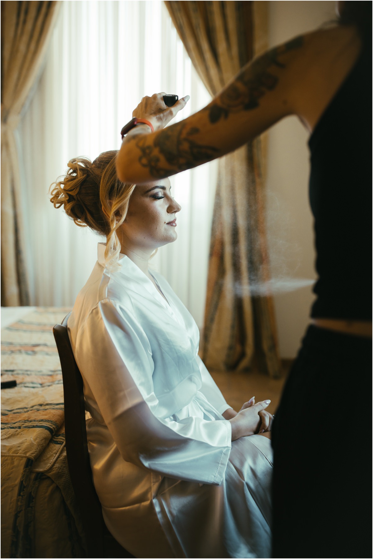 Bride Hairstyle Being Finished Hairstylist spraying the updo of bride Miriana, sitting in a satin robe by the window