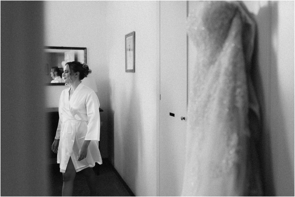 The Wedding Dress Waiting, Black and White Black and white photo of the lace wedding dress hanging on a door while bride Miriana walks in the room