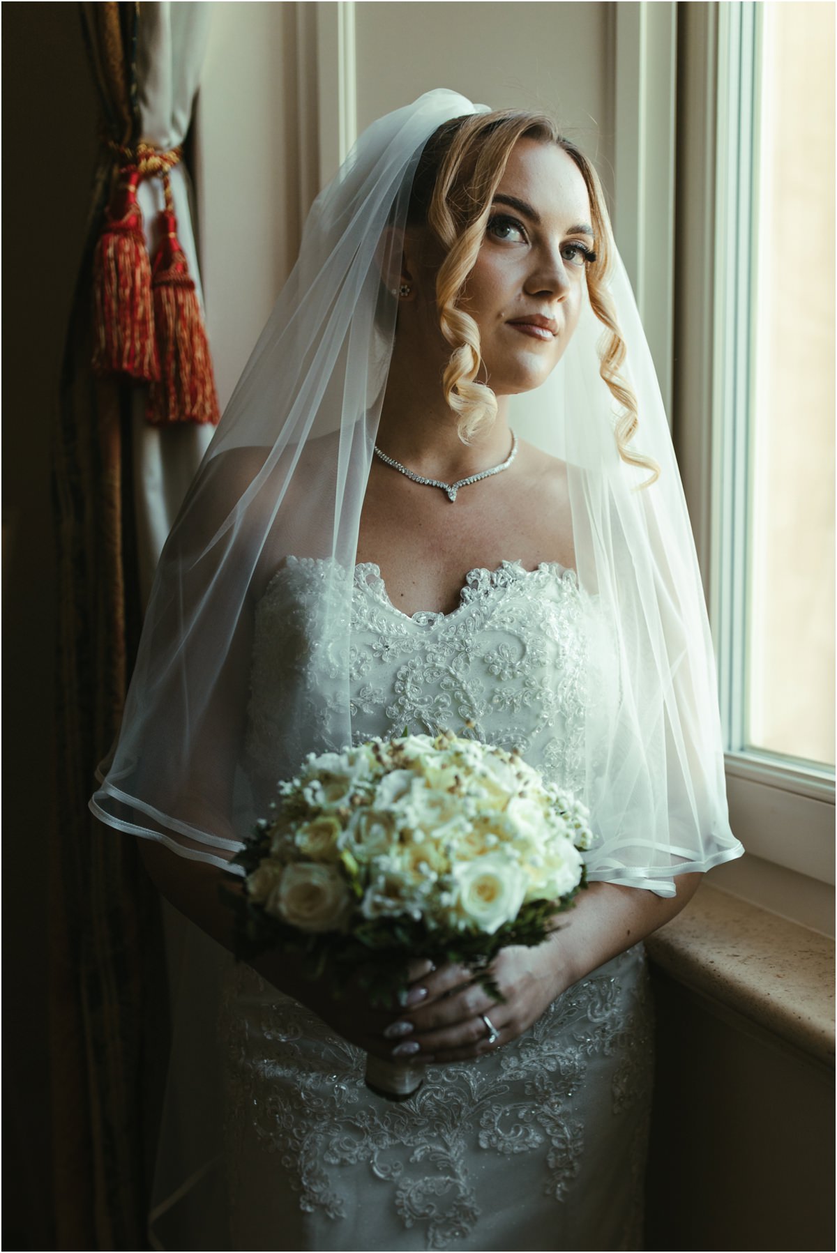 Bride Portrait by the Window Portrait of bride Miriana with veil and white rose bouquet, looking out of the window in natural light