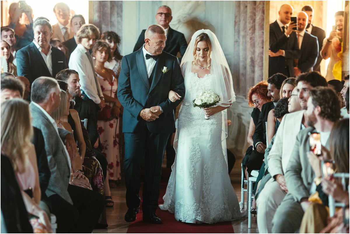 Bride Miriana Entering the Ceremony Bride Miriana walking in on the arm of a man in a tuxedo while guests stand and film at Villa Grazioli