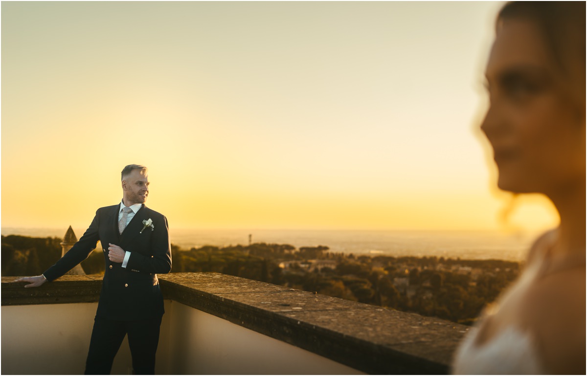 Angus Waiting for Miriana on the Terrace Groom Angus leaning on the terrace wall at sunset, with Miriana out of focus in the foreground