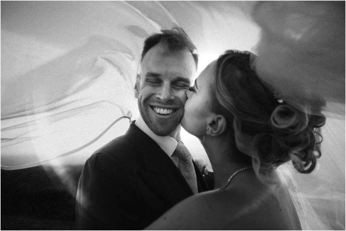 A Kiss Under the Veil, Black and White Black and white photo of Miriana kissing laughing Angus on the cheek, her veil blowing in the wind