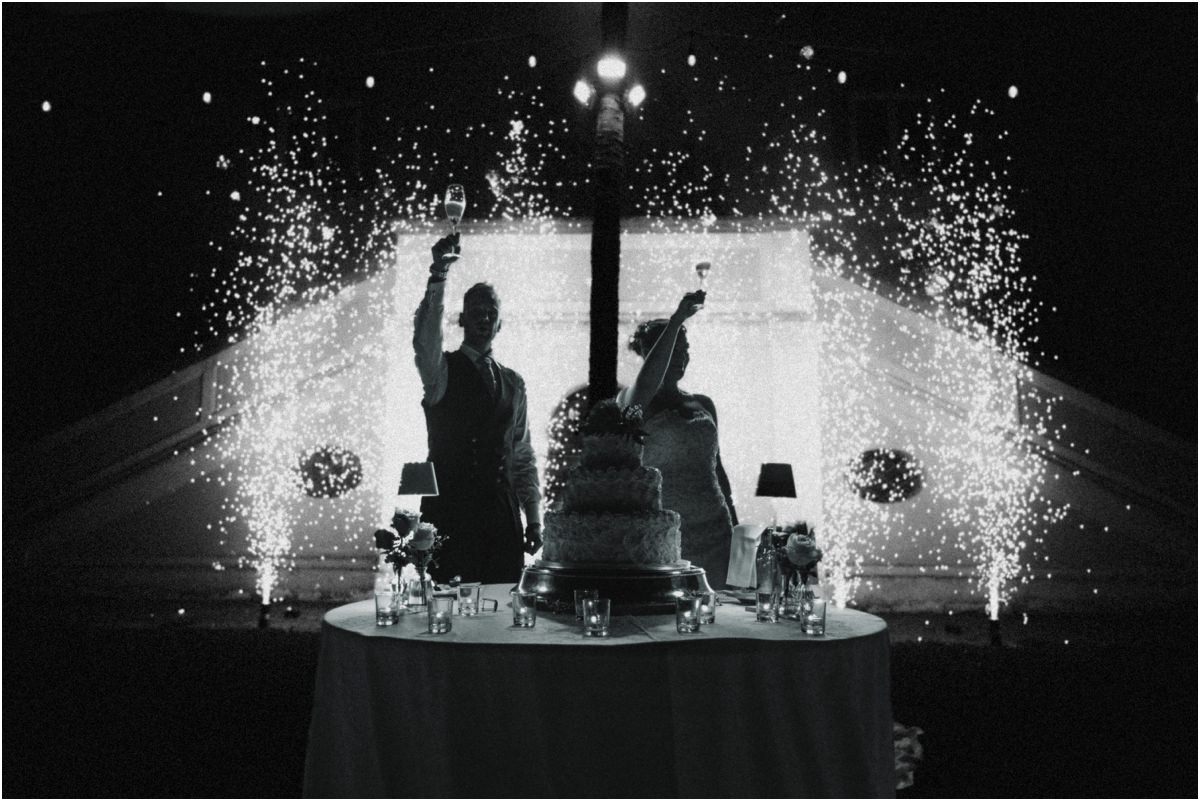 Cake Toast with Sparklers, Black and White Black and white silhouette of Miriana and Angus raising their glasses by the wedding cake with sparkler fountains
