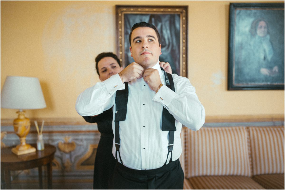 Tying the Bow Tie A family member tying groom Alessio's bow tie in a yellow room with old portraits at Villa Grazioli