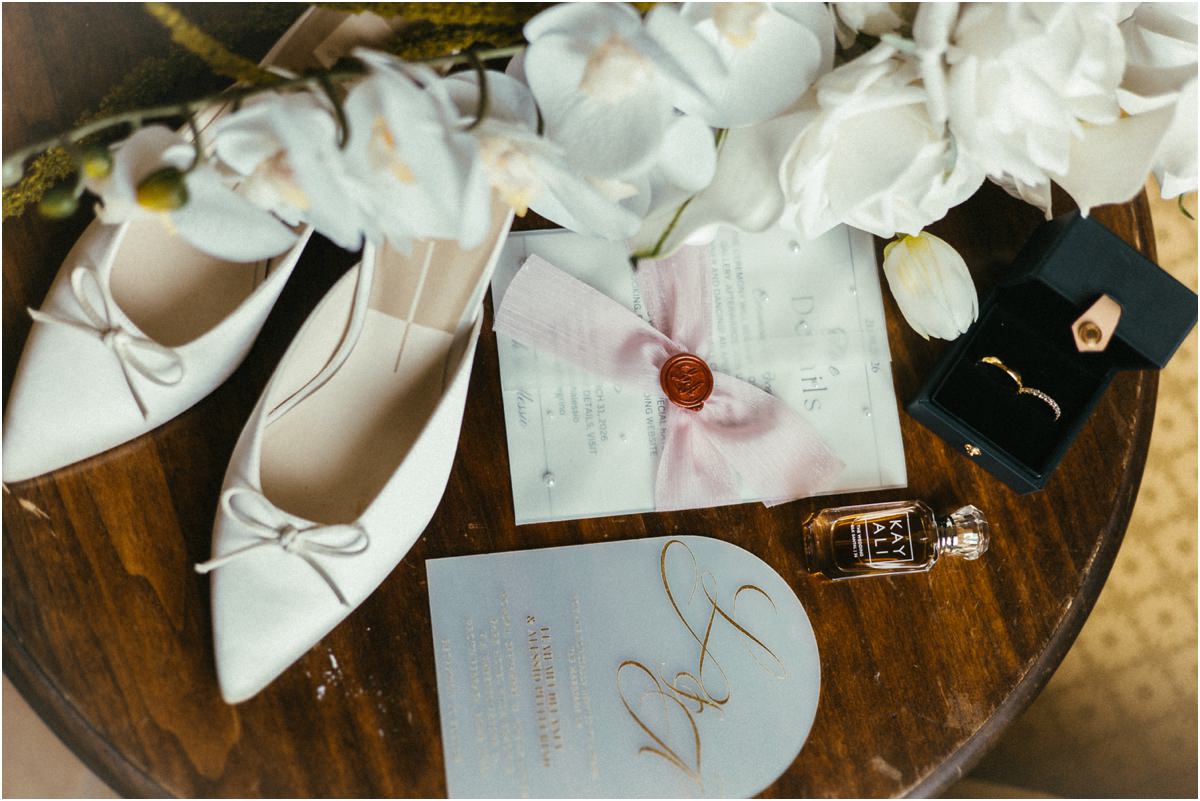 Bridal Details: Shoes, Rings and Invitation Bridal details for Leah: white bow flats, wedding rings, wax sealed invitation, perfume and white orchids on a wooden table