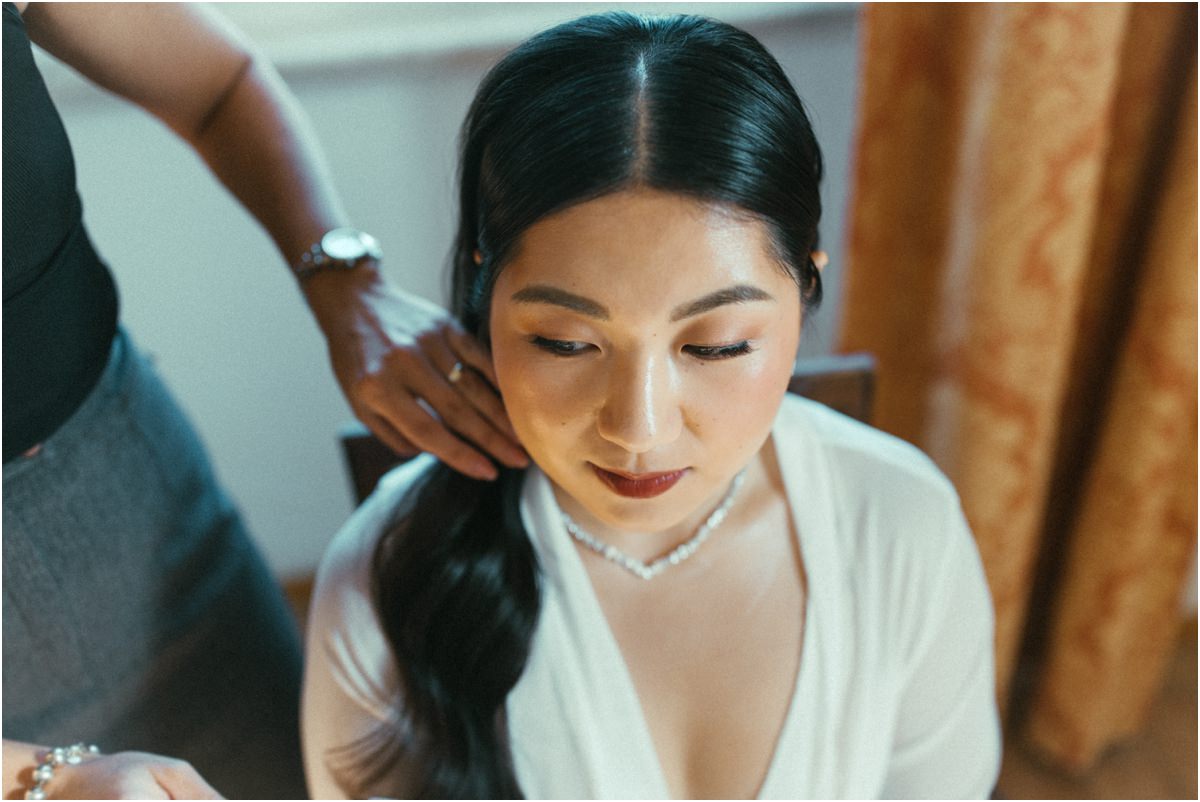 Final Touches to the Hair Bride Leah looking down with red lipstick and a diamond necklace while her hair is finished