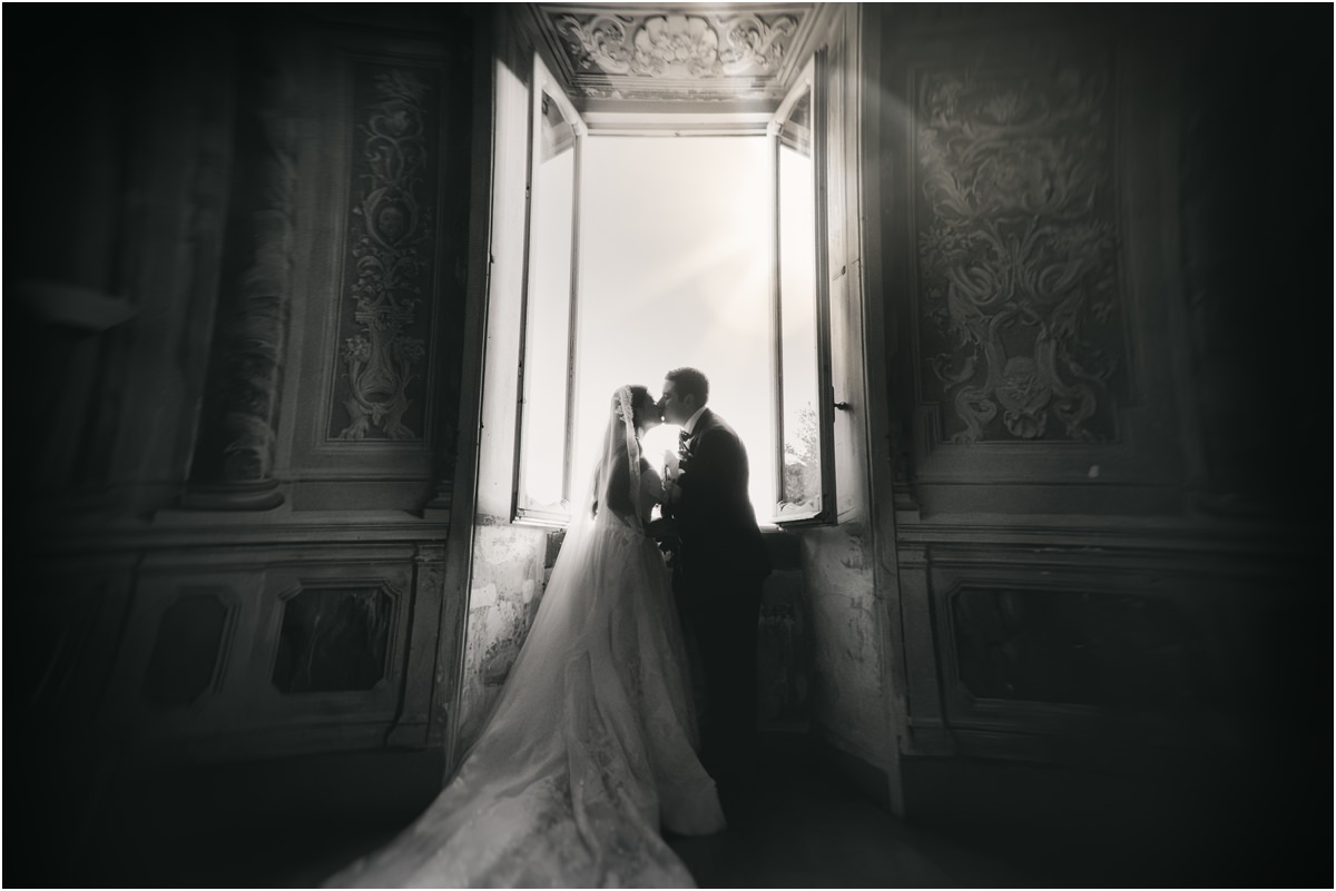 Kiss by the Open Window Black and white photo of Alessio and Leah kissing in front of a tall open window, her long veil spread on the floor