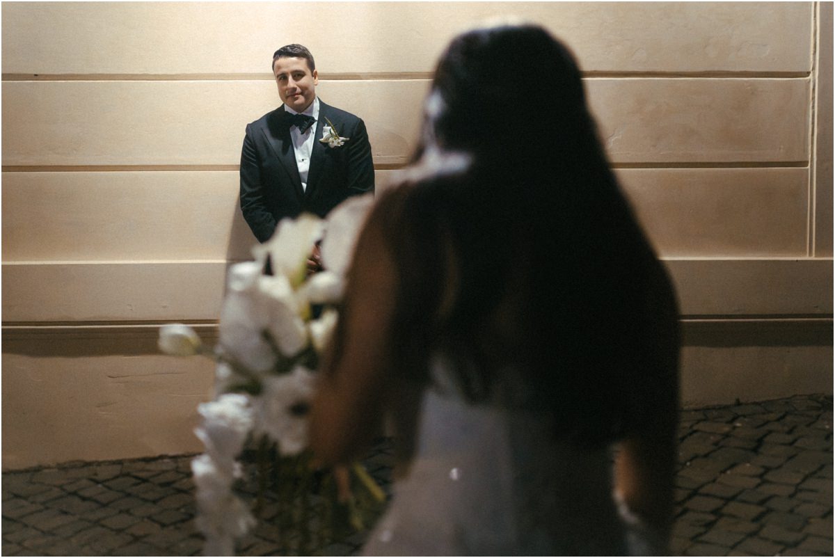 Waiting for Leah Groom Alessio leaning against a wall in the evening while Leah walks towards him with her bouquet