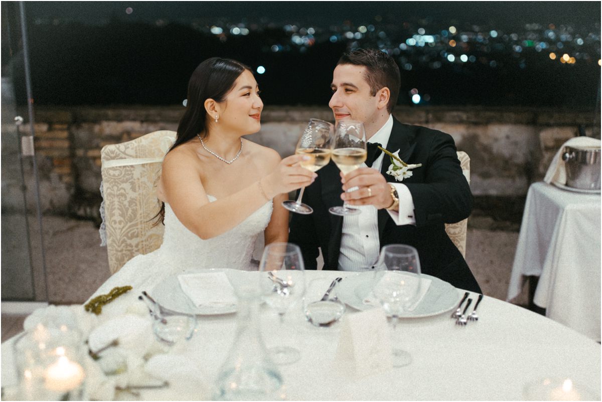 A Toast at the Couple's Table Alessio and Leah toasting with white wine at their table, city lights in the distance