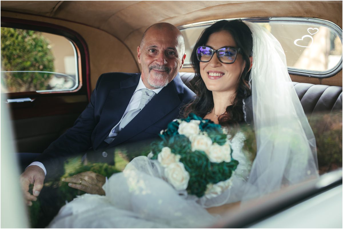 Verso la chiesa Martina sorridente con velo e bouquet seduta nell'auto d'epoca accanto al signore in abito blu