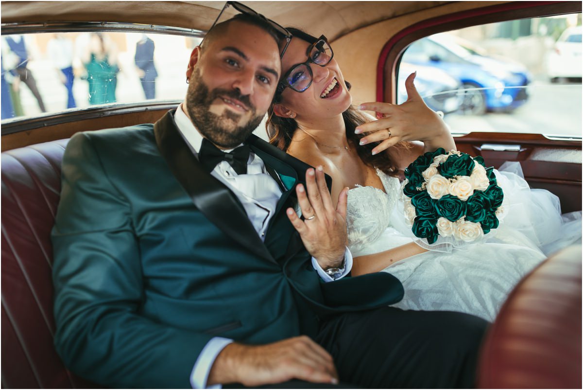 Sposati Martina e Davide ridono nell'auto d'epoca mostrando le fedi, con il bouquet verde e bianco