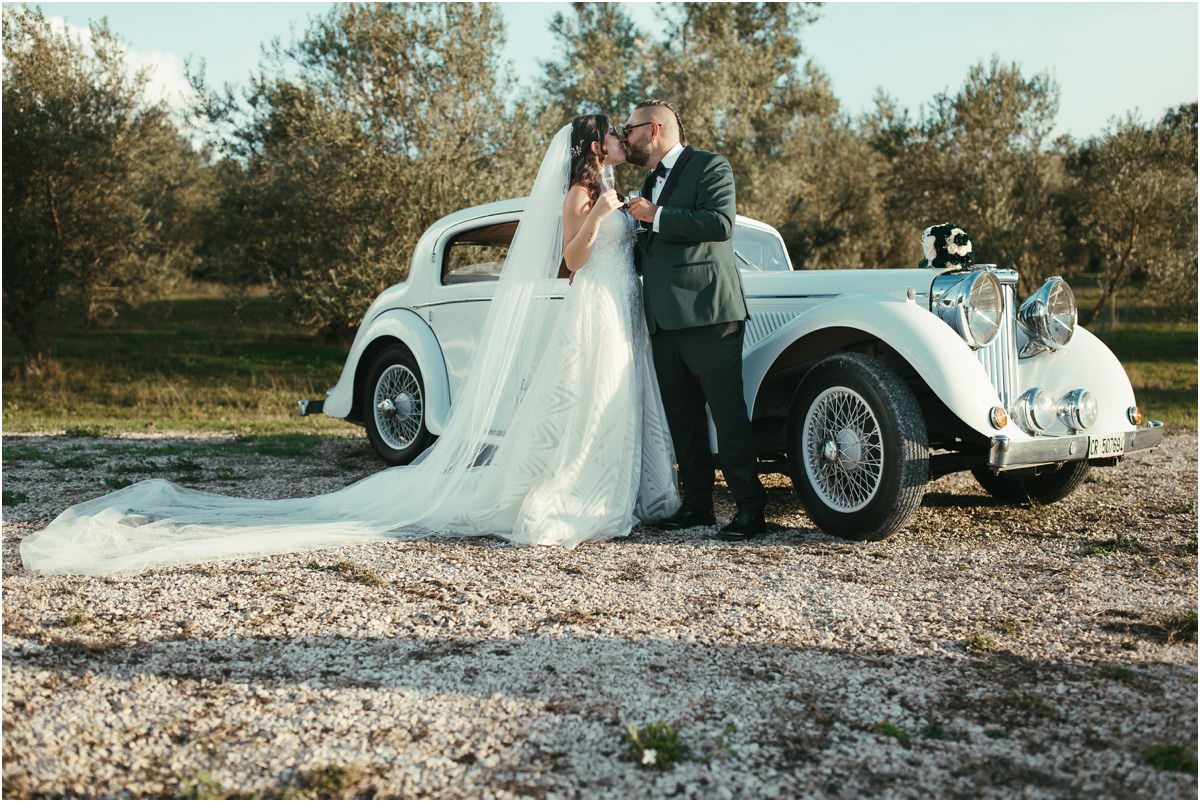 Un bacio accanto all'auto Martina e Davide si baciano appoggiati all'auto d'epoca bianca tra gli ulivi di Le Jardin Potager, con il lungo velo sulla ghiaia